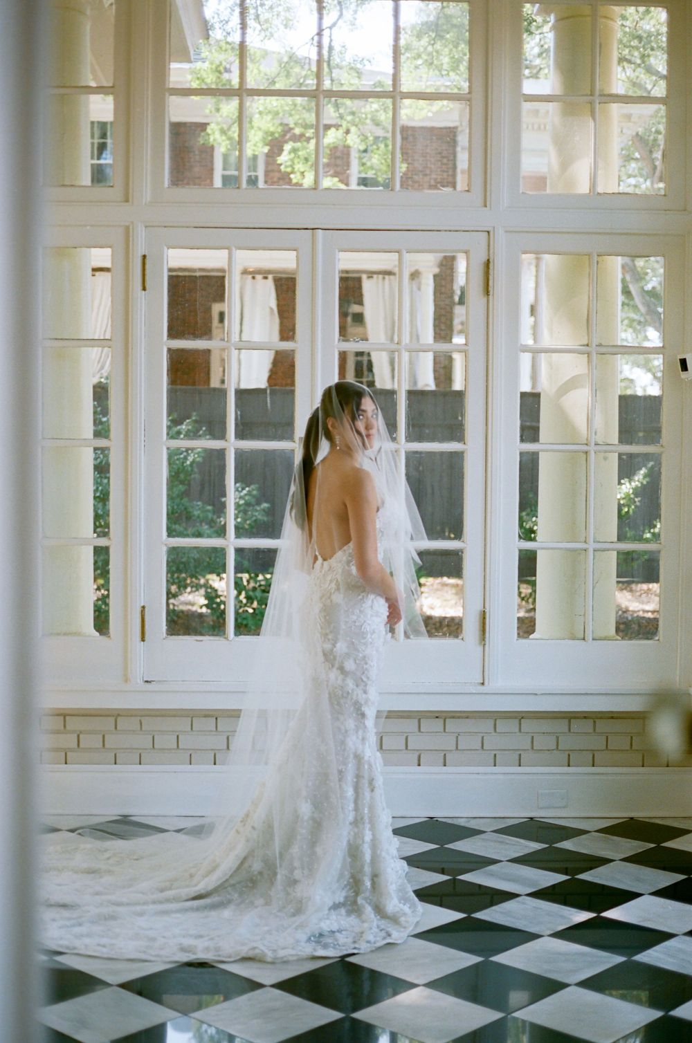Wedding gown with cathedral train on black and white checkered floor in sunlit room.