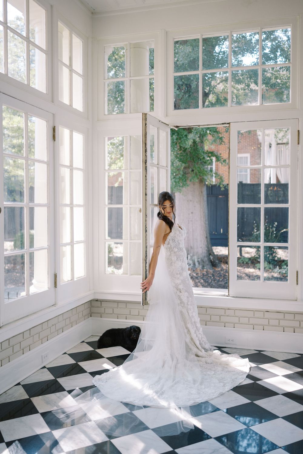 A bride in a flowing white wedding gown stands in a sunlit room with black and white checkered floors.