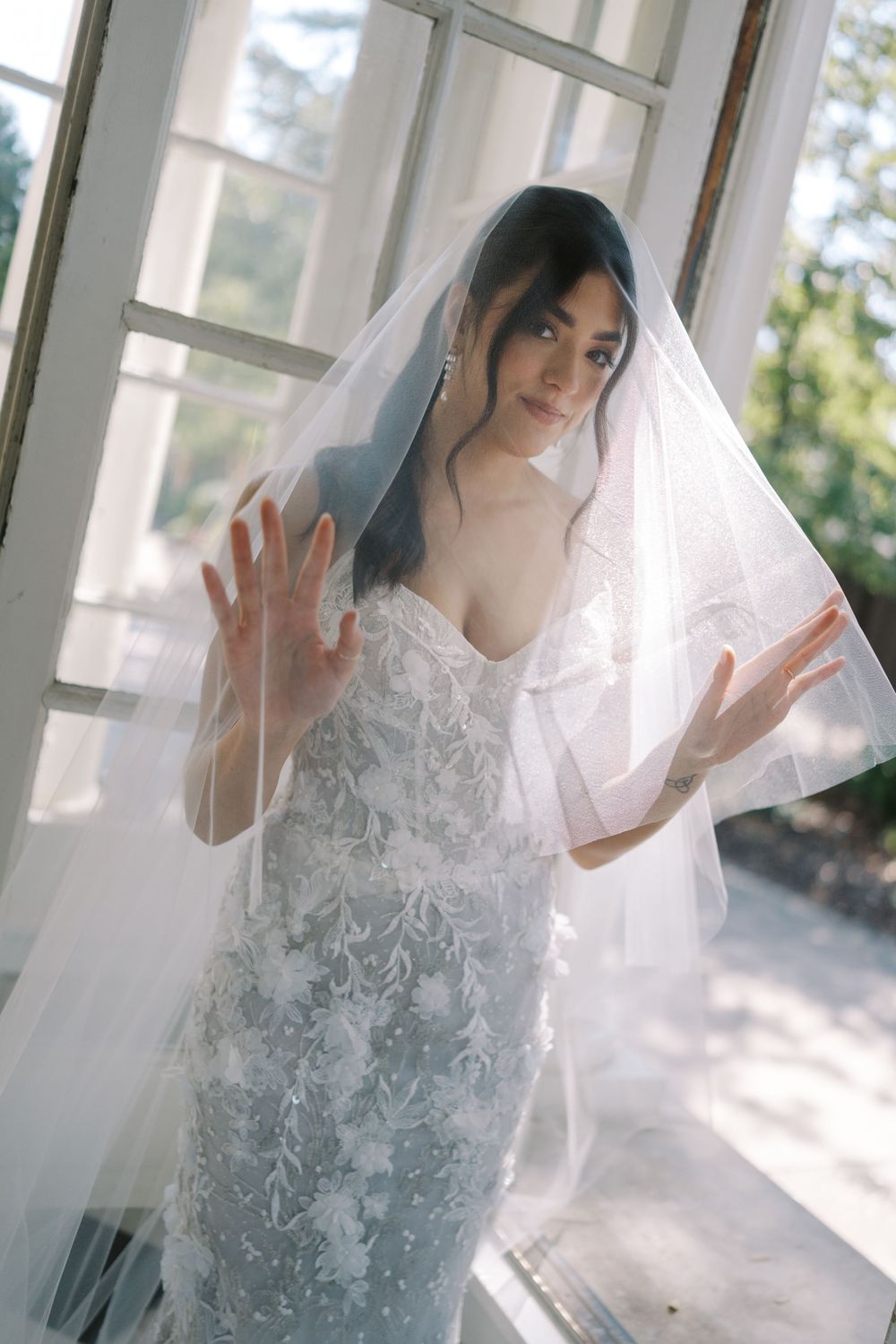 Ethereal bridal portrait with flowing veil in a sunlit room with white walls.