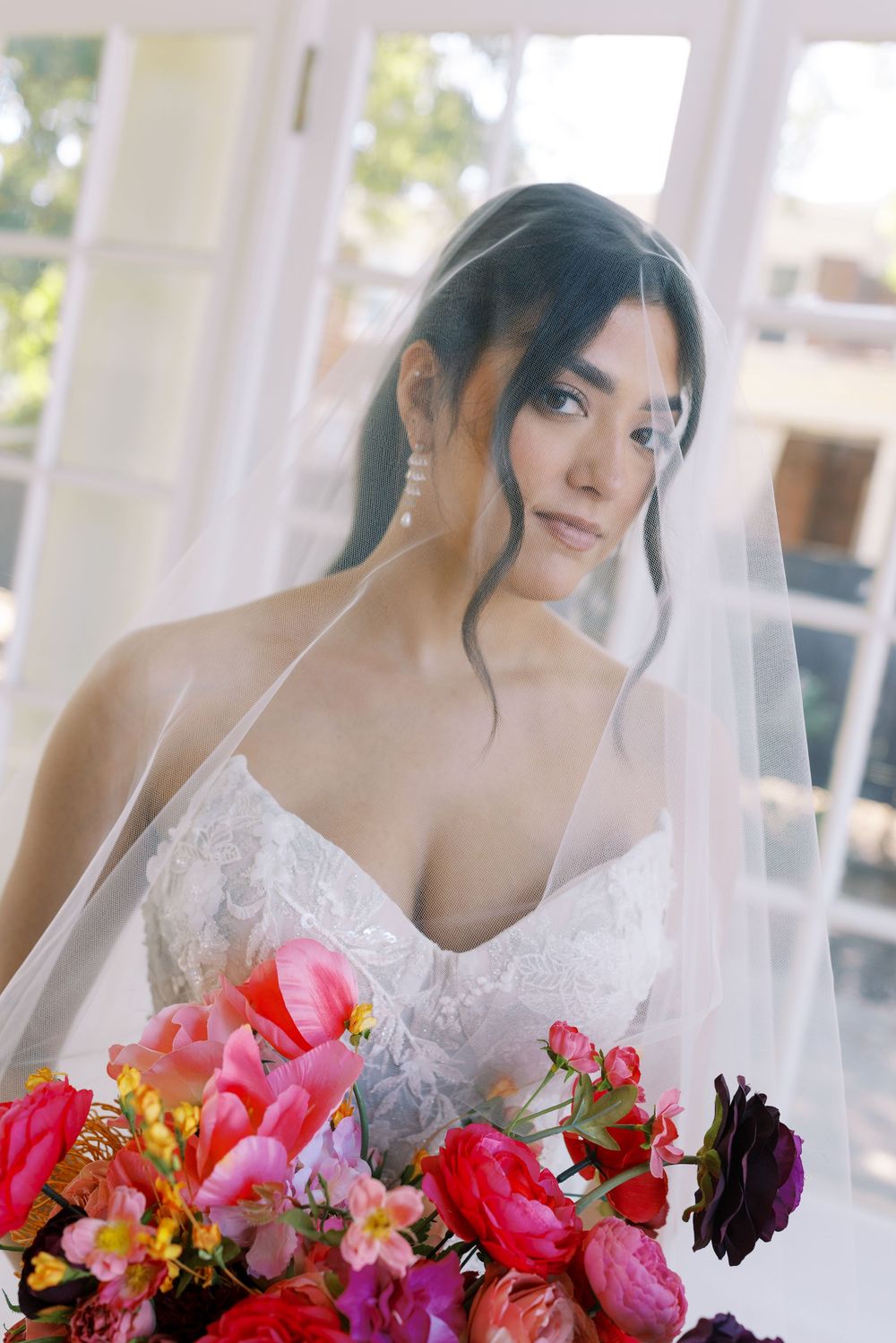 Elegant bride in lace gown holds vibrant pink and coral bouquet against white curtained backdrop.