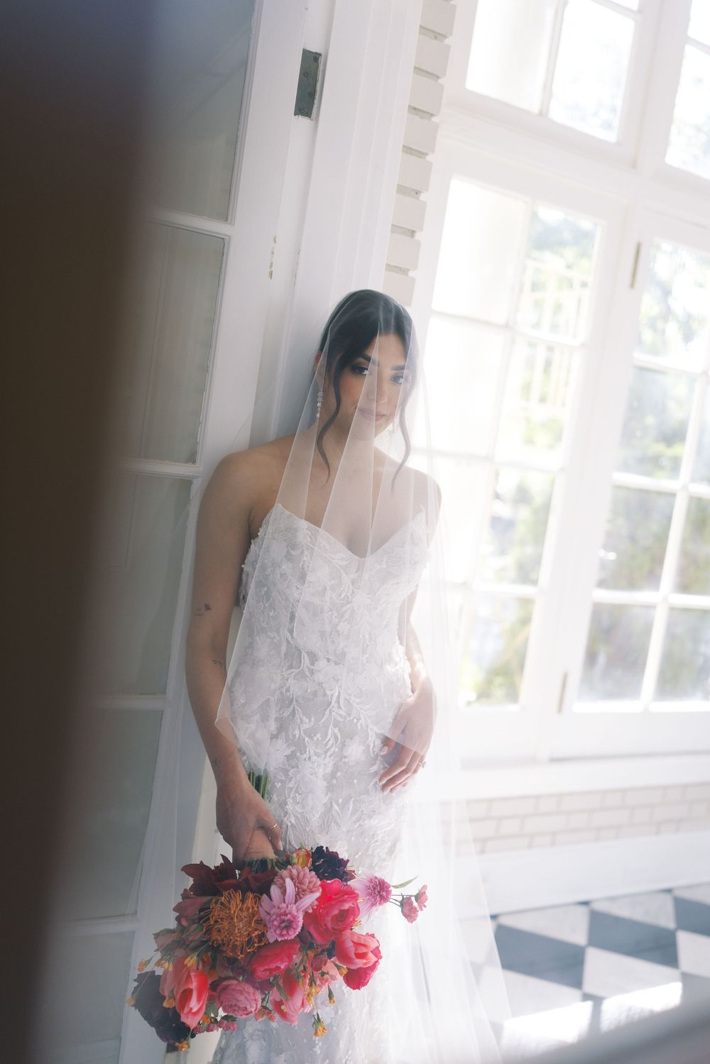 Bride in white lace wedding dress holds vibrant red and pink bouquet while standing near bright window.
