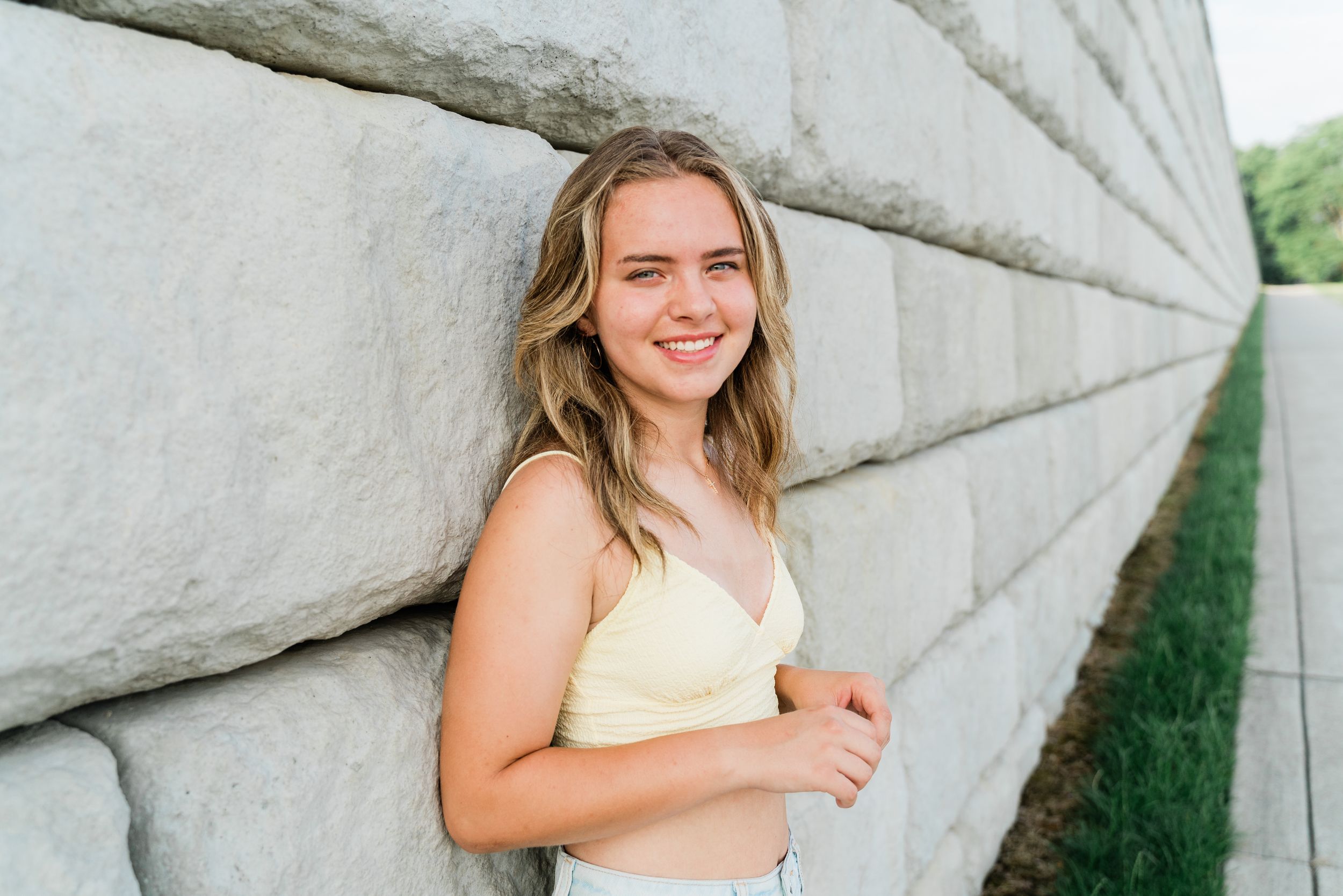 A person in a light yellow top smiles while standing against a textured concrete wall outdoors.