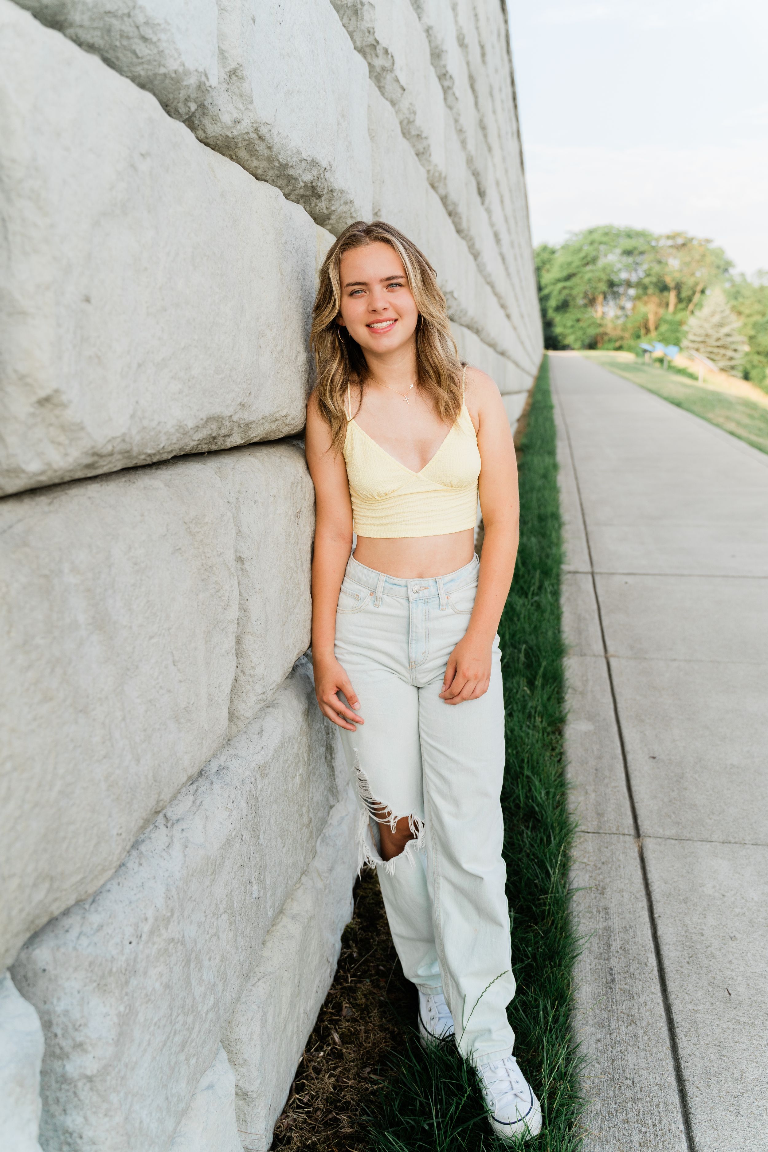 A person in a yellow crop top and ripped jeans poses casually against a concrete wall on a sunny day.