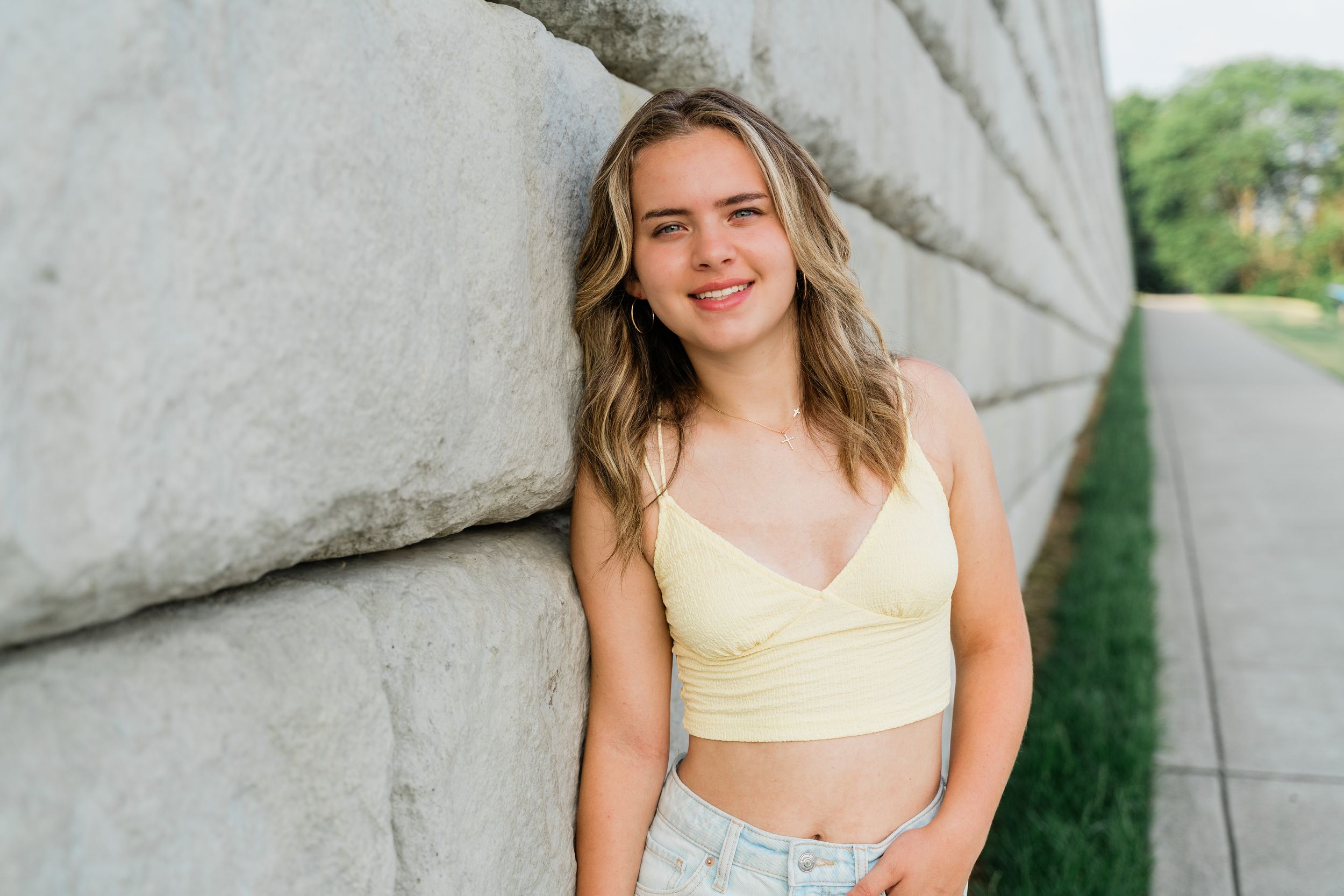 A person in a yellow top poses outdoors against a concrete wall backdrop during a sunny day.