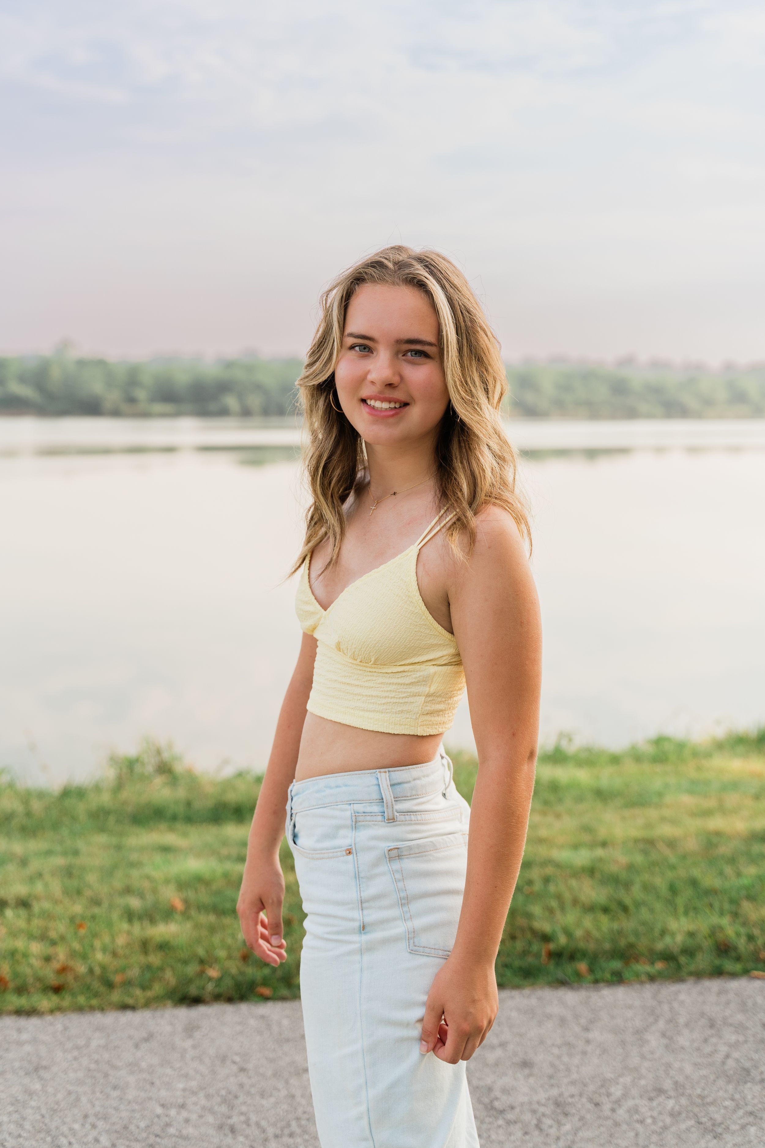 A person in a yellow crop top and light jeans poses outdoors by a lake on a sunny summer day.