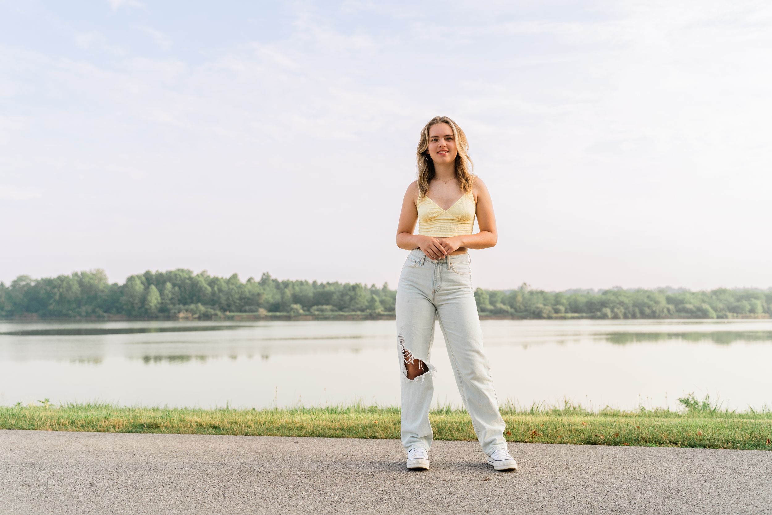Woman in white dress poses in a scenic outdoor setting by a lake during golden hour.