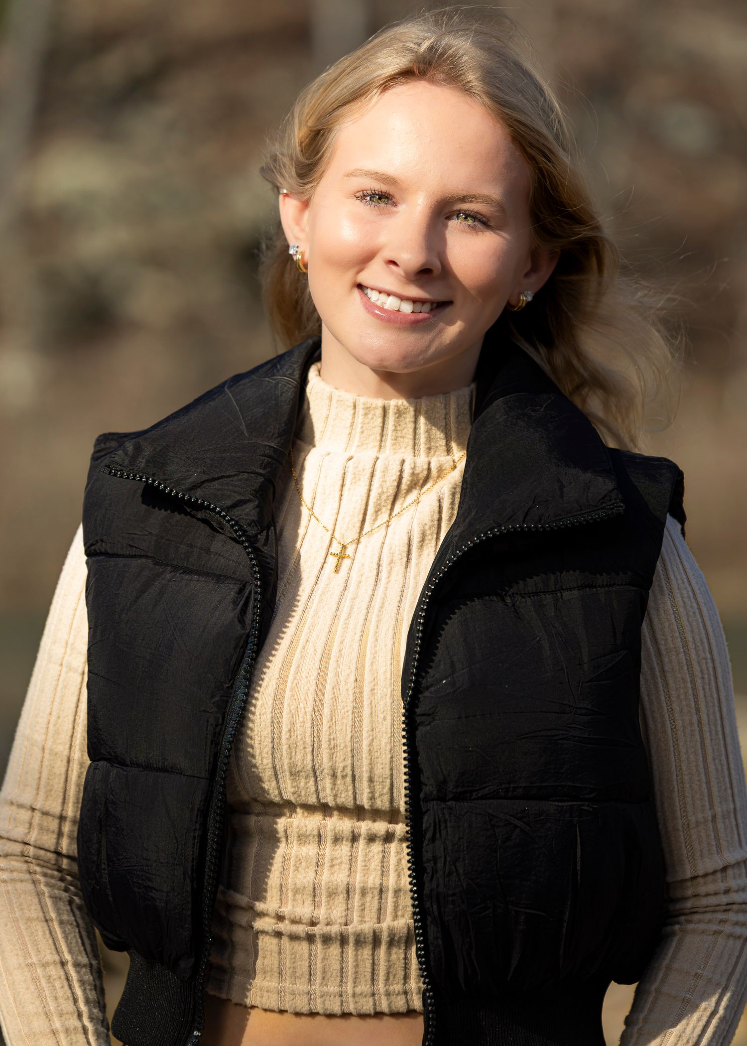 A person wearing a black puffer vest and cream ribbed sweater smiles warmly in an outdoor setting.