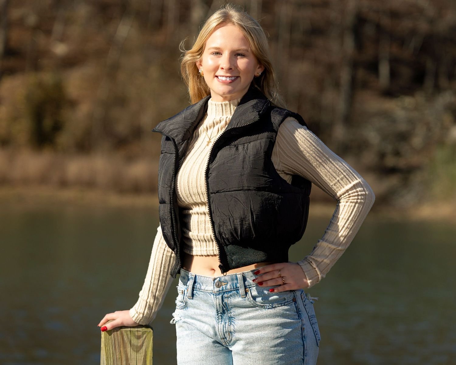 Person in a black puffer vest and cream sweater poses near a lake on a sunny autumn day.