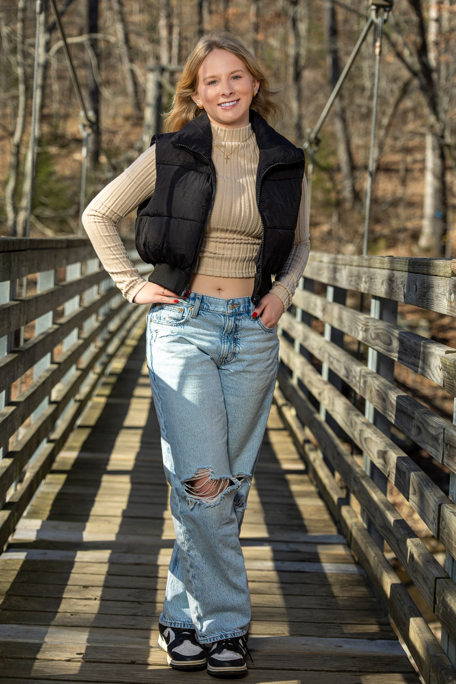 A person in a cream sweater, black puffer vest and ripped jeans poses on a wooden bridge in a forest setting.