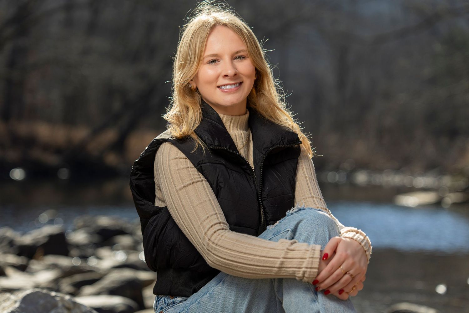 Person in black vest and jeans sitting casually by waterfront during autumn season.