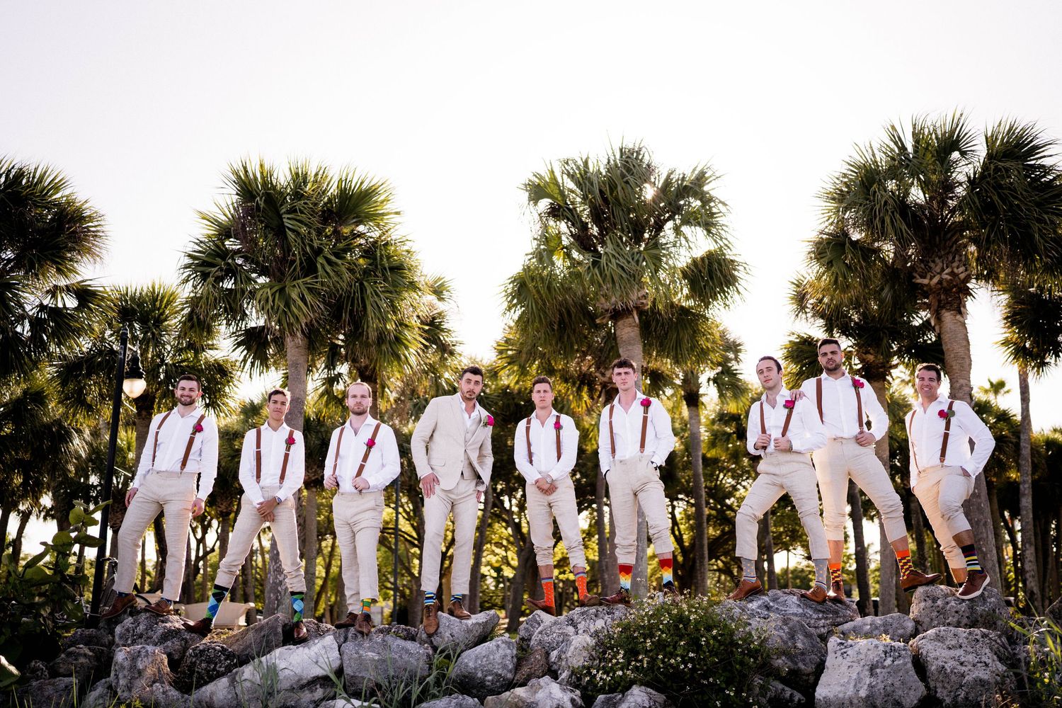 Wedding party in white shirts and suspenders poses together against palm trees at sunset.
