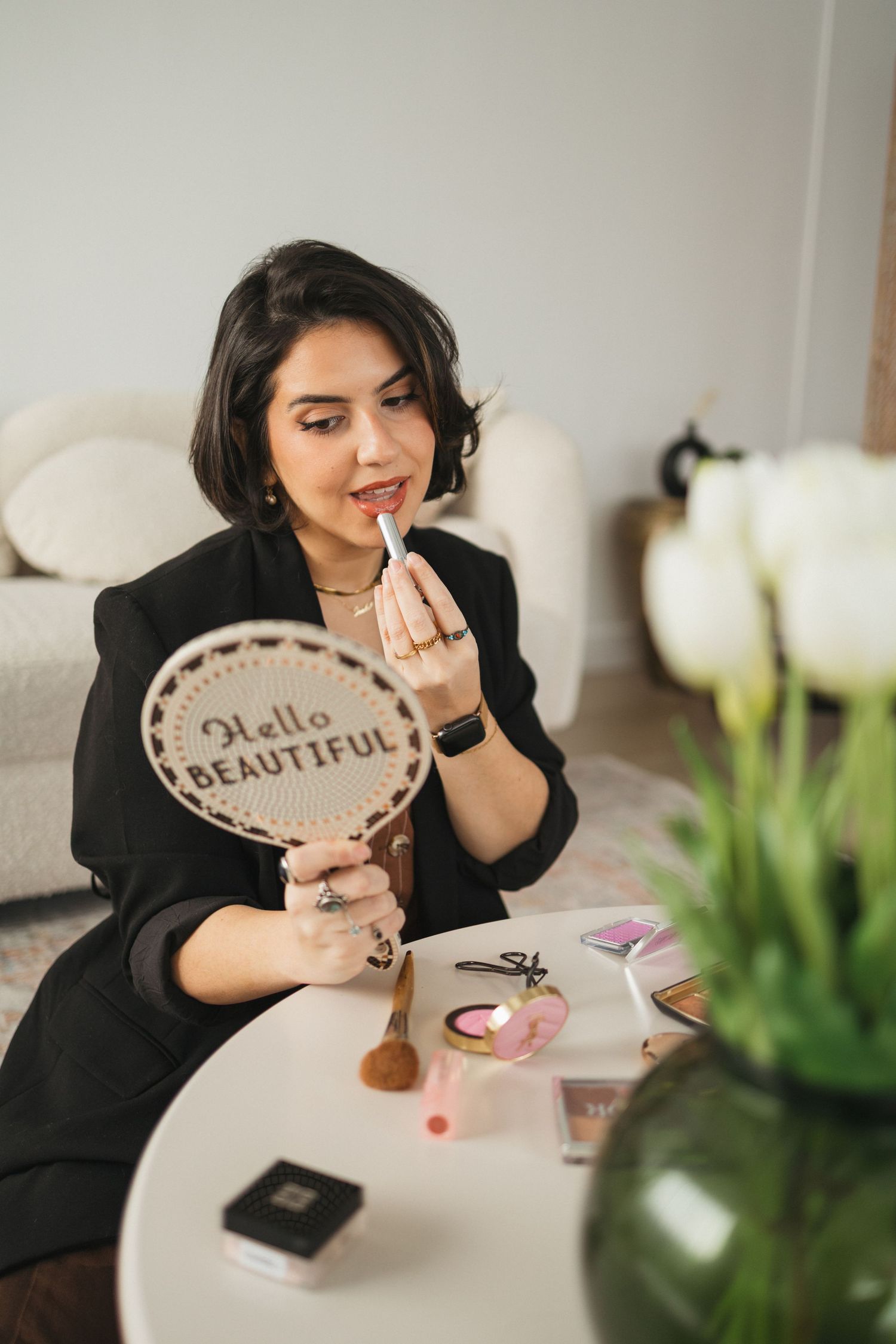 Person applying lipstick while holding a mirror that says 'Hello Beautiful' with flowers in background.