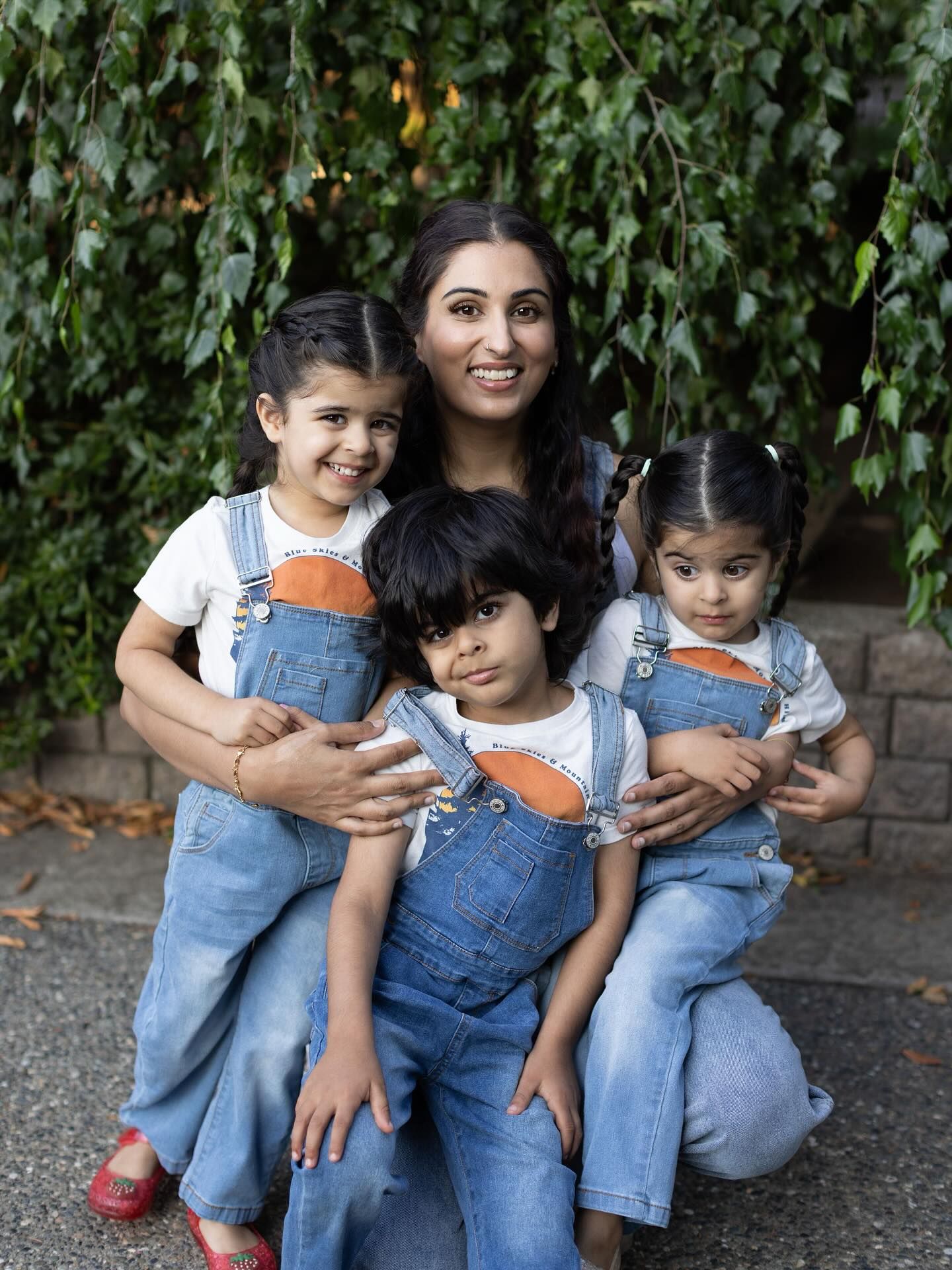 A family wearing matching denim overalls sits together outdoors against a leafy green backdrop.