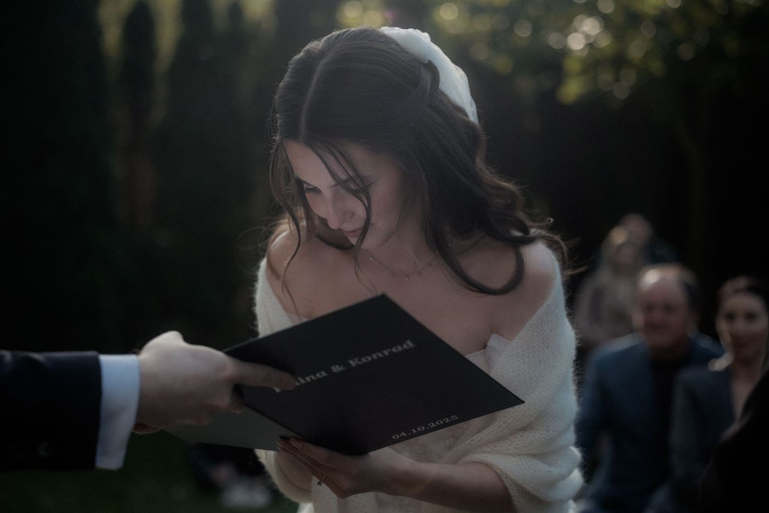 A person in white holds a book during an outdoor ceremony with guests visible in the background.