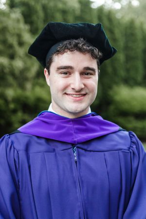 Graduate wearing blue academic regalia with purple hood smiling during commencement ceremony outdoors.
