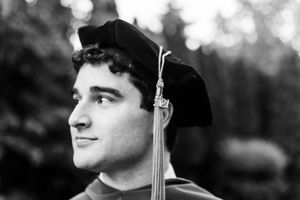 Graduate in cap and gown looking thoughtfully into the distance during outdoor commencement ceremony.