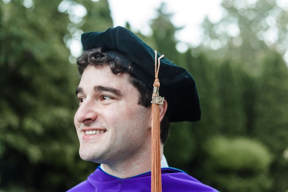 Graduate in purple and black regalia smiling during outdoor commencement ceremony with trees in background.