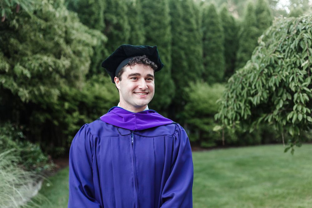 Graduate in blue cap and gown stands outdoors in front of green trees during graduation photo session.