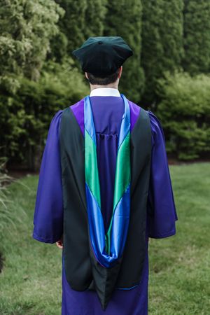 Academic graduation gown shown from behind with blue and teal stole against green outdoor background.