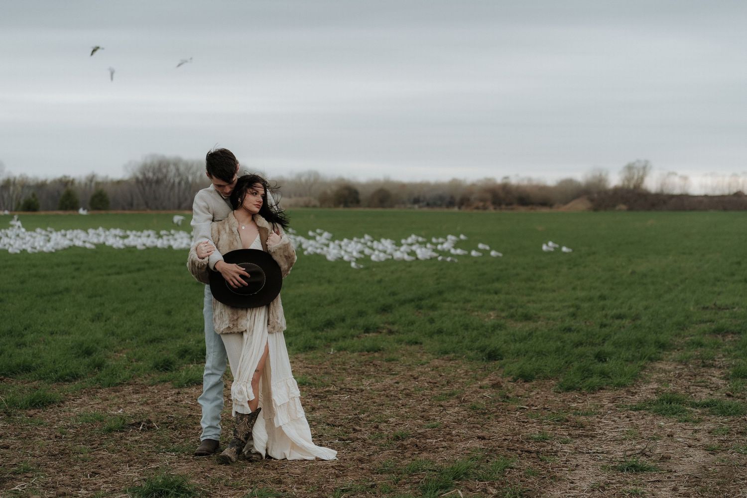 A romantic couple in light-colored outfits embrace tenderly in a golden wheat field on an overcast day.
