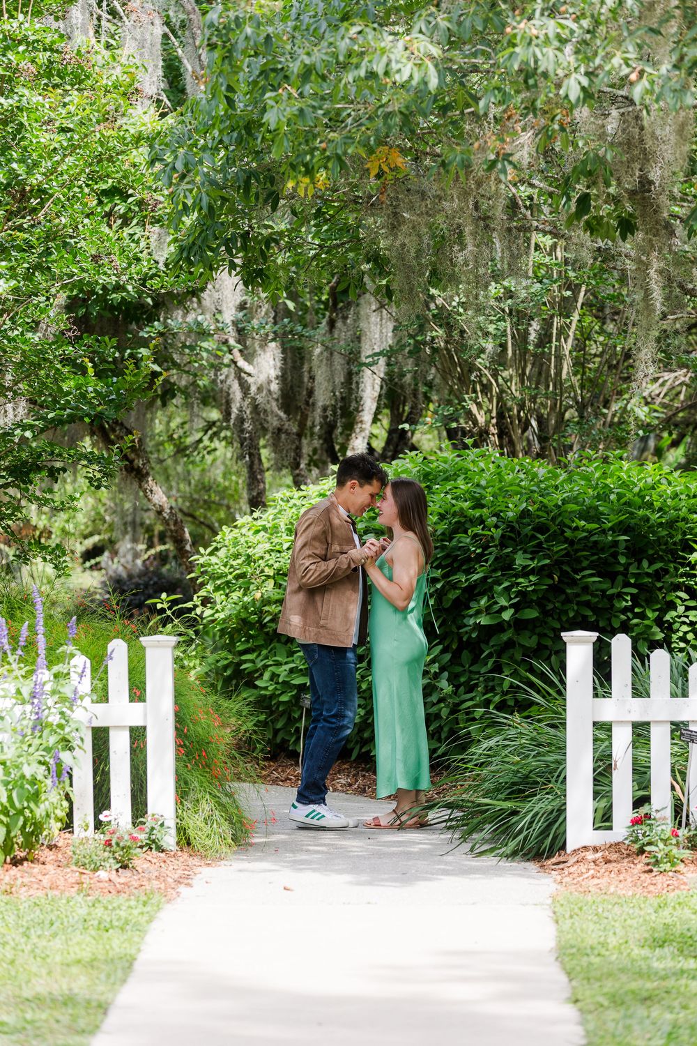 A couple shares an intimate moment on a garden path framed by Spanish moss and white picket fence posts.