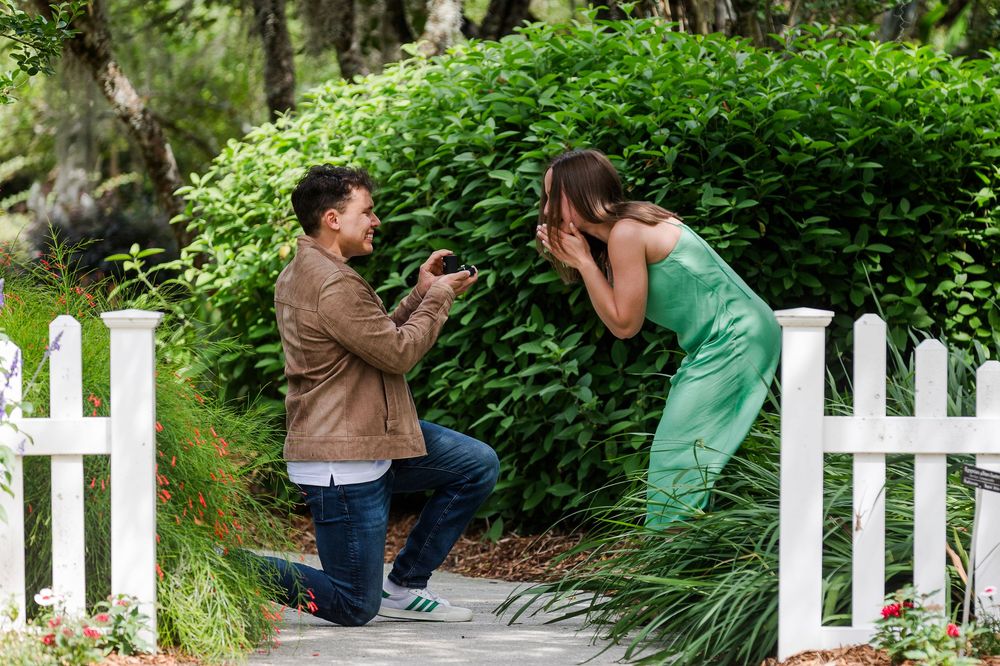 A romantic proposal moment captured in a lush garden setting with a white picket fence in the background.
