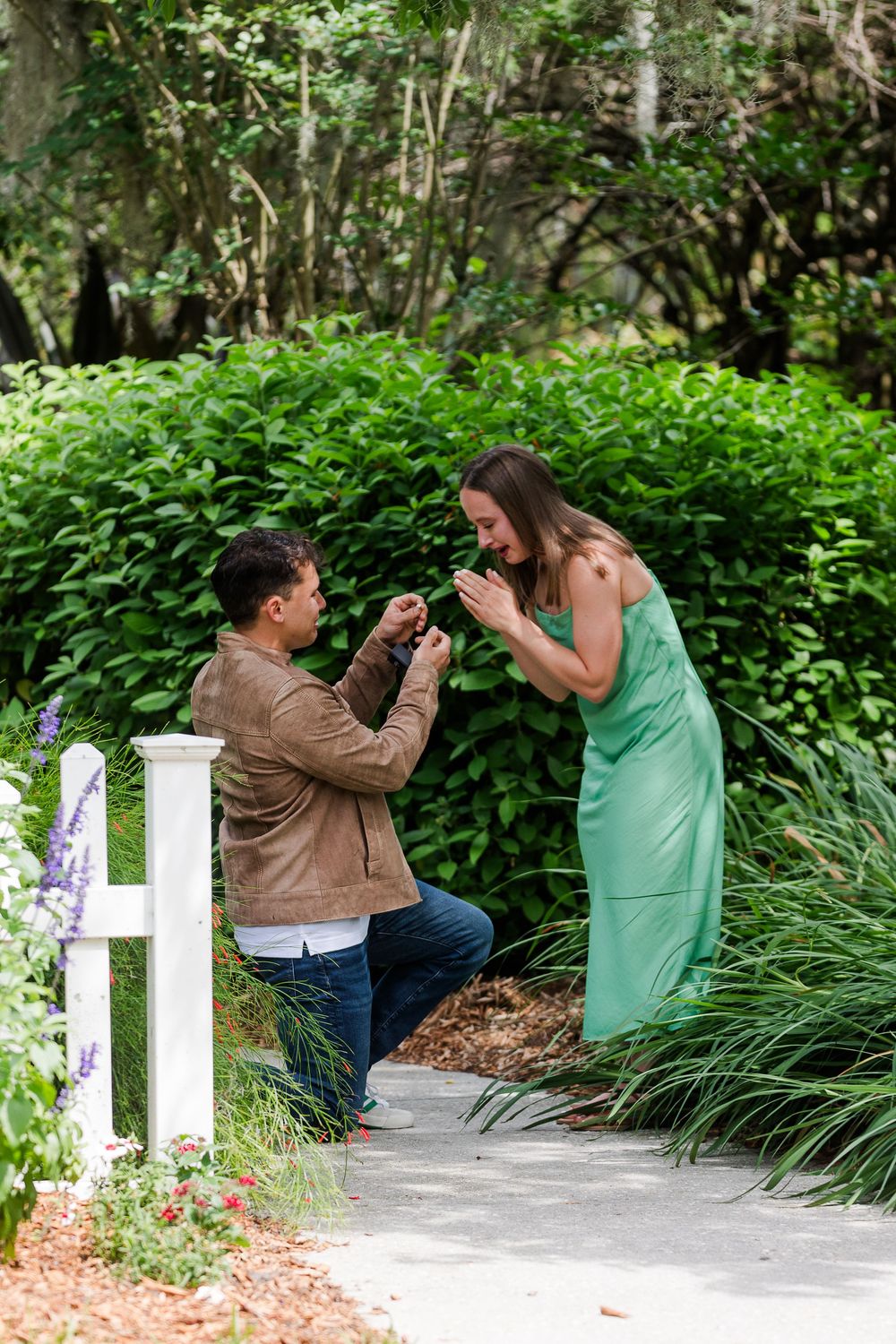 A romantic proposal moment captured on a garden path surrounded by lush greenery and a white fence.