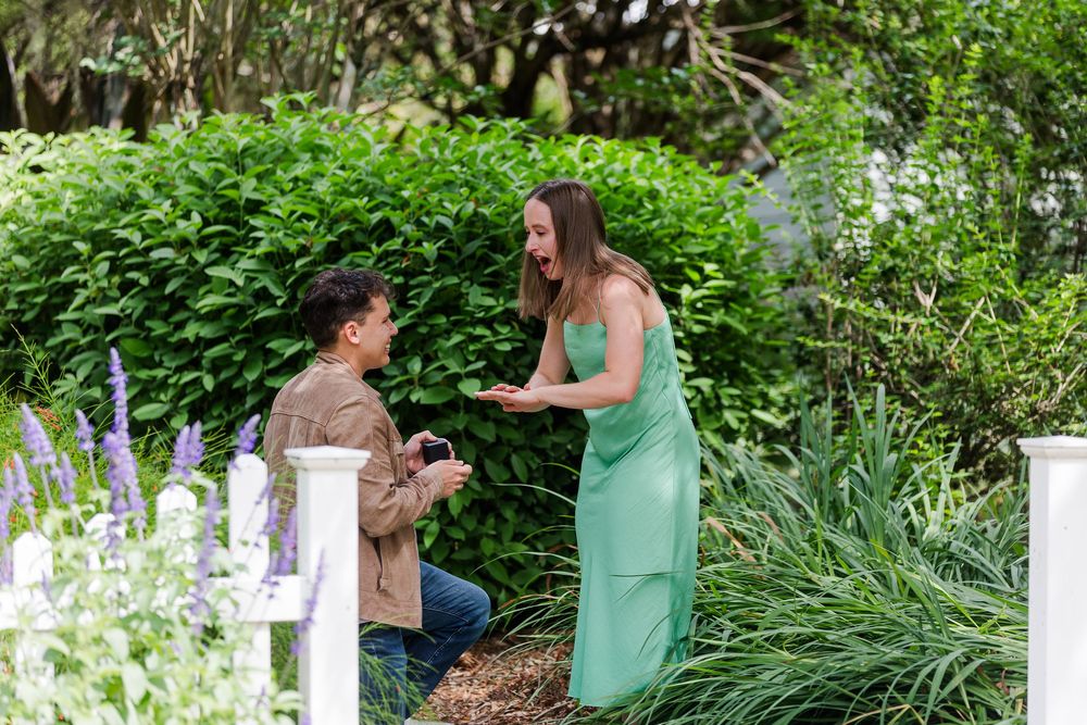 A couple embraces in a romantic moment outdoors surrounded by lush green plants and white garden fencing.