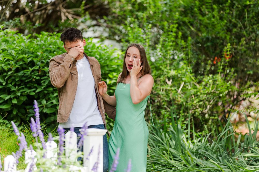 A couple walks through a lush garden path filled with blooming flowers and greenery on a sunny summer day.