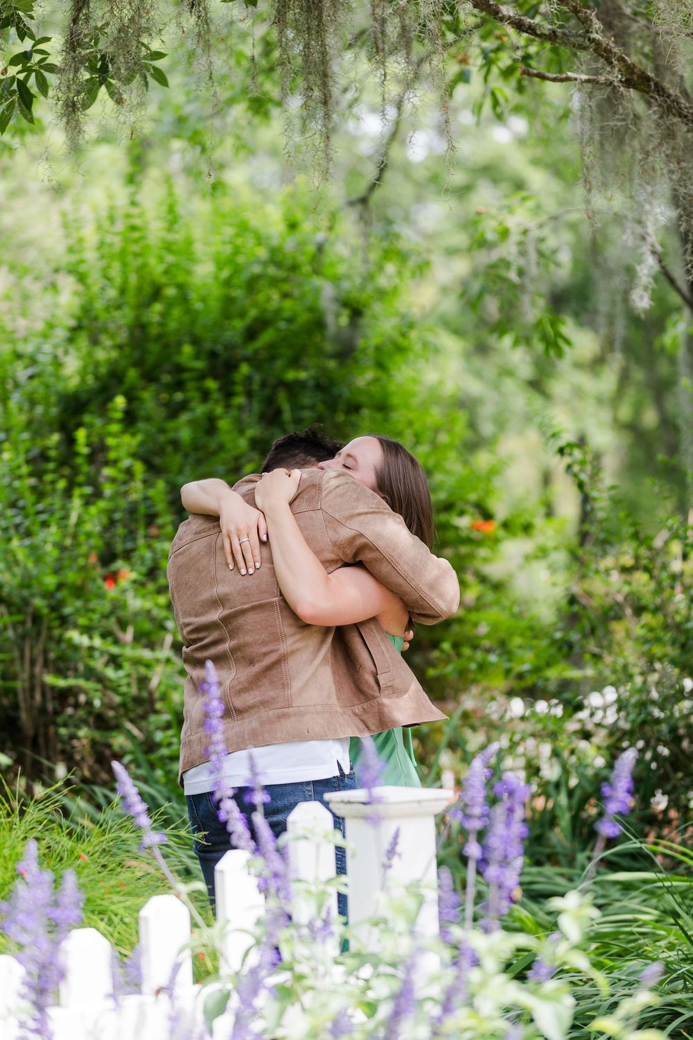 Two people share a tender moment in a lush garden with purple flowers and Spanish moss.
