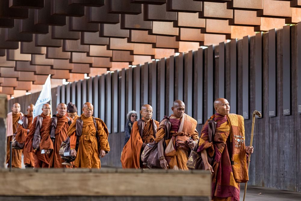 Buddhist monks in traditional orange robes walk in a line through a temple corridor during a ceremony.