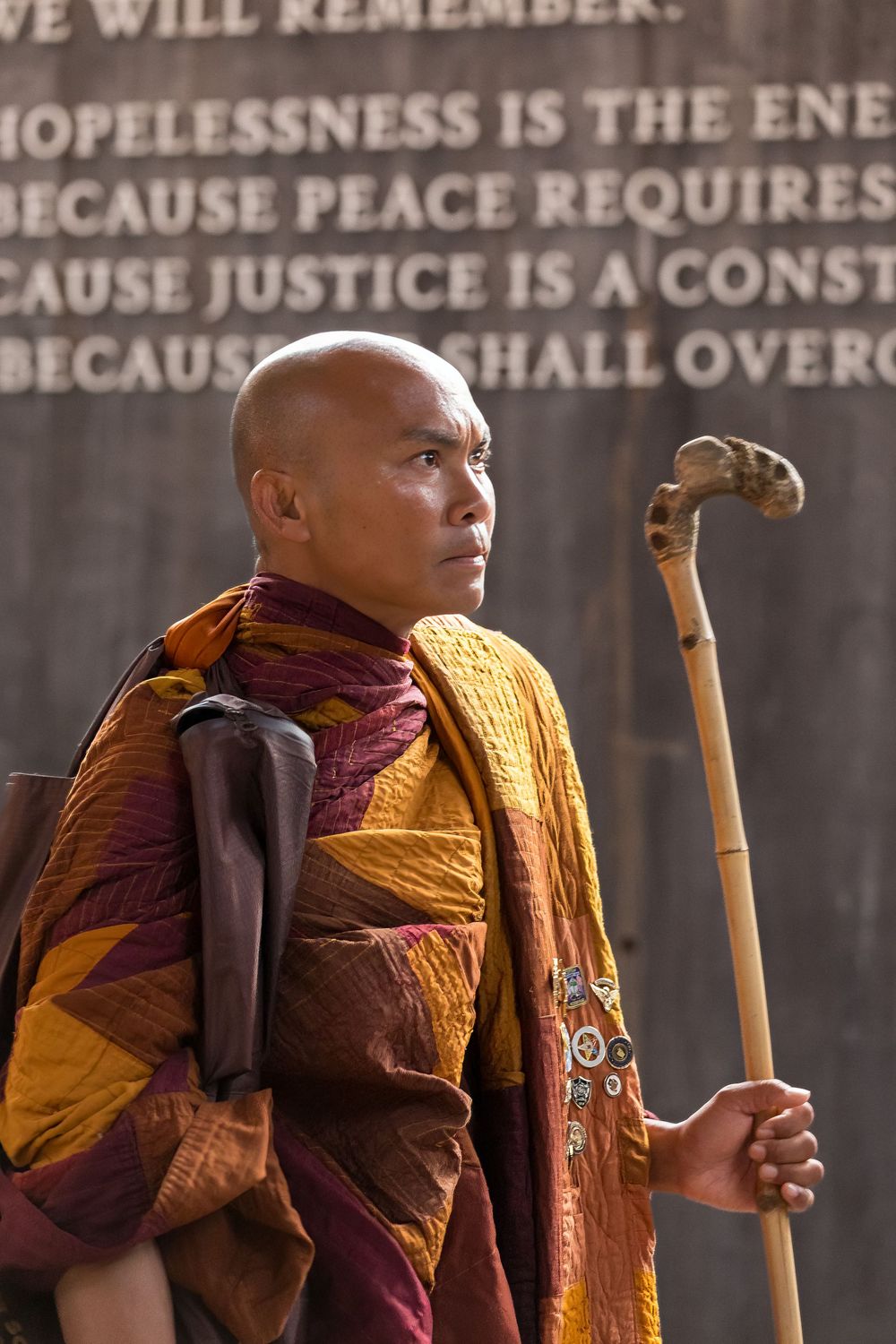 Buddhist monk in traditional orange and burgundy robes holds a wooden staff against a wall with text.
