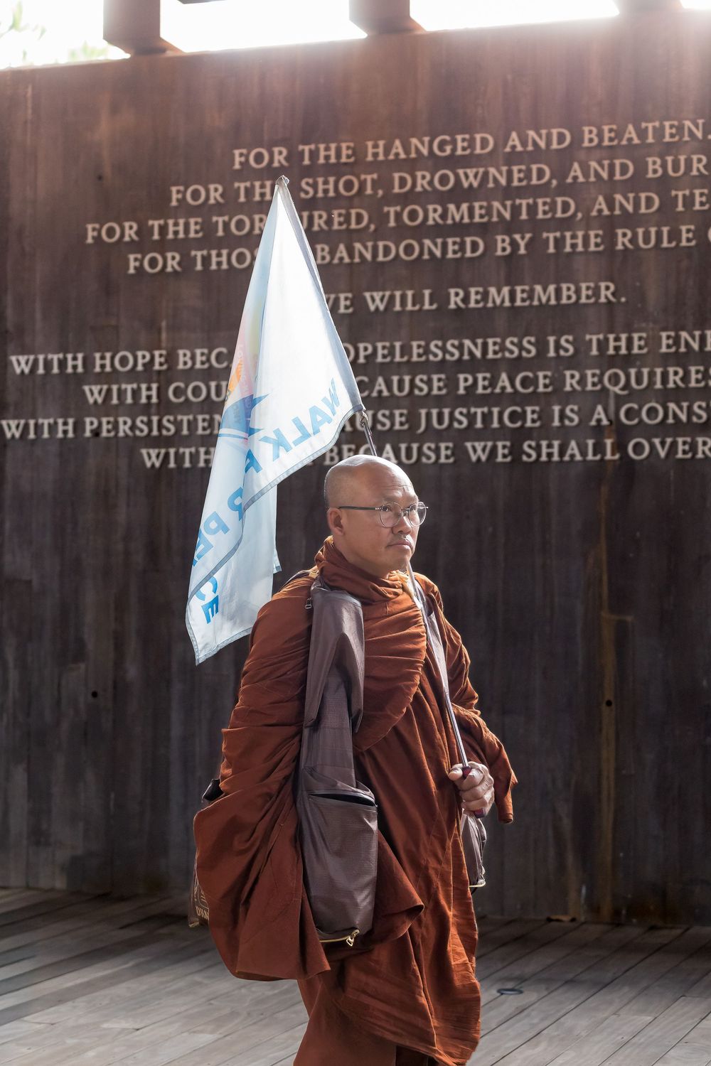 A Buddhist monk in traditional brown robes stands in front of a memorial wall with text about remembrance and justice.