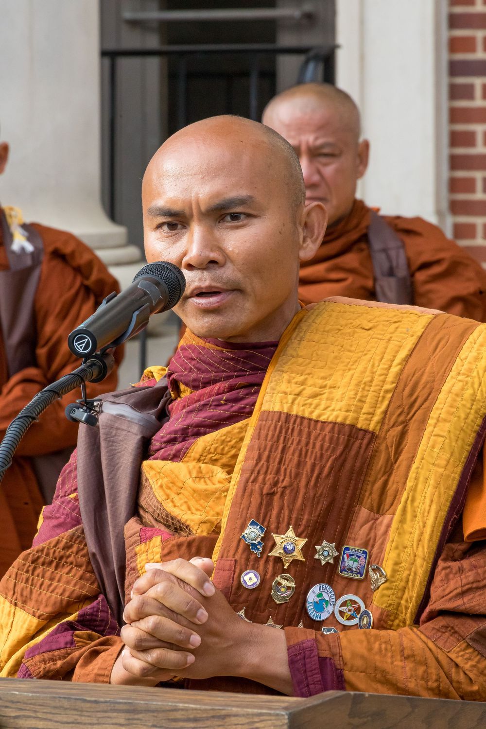 Buddhist monk in saffron robes holds a microphone while speaking during a ceremony.