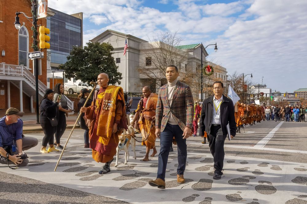 Buddhist monks and civilians walk together across a crosswalk in an urban setting under blue skies.
