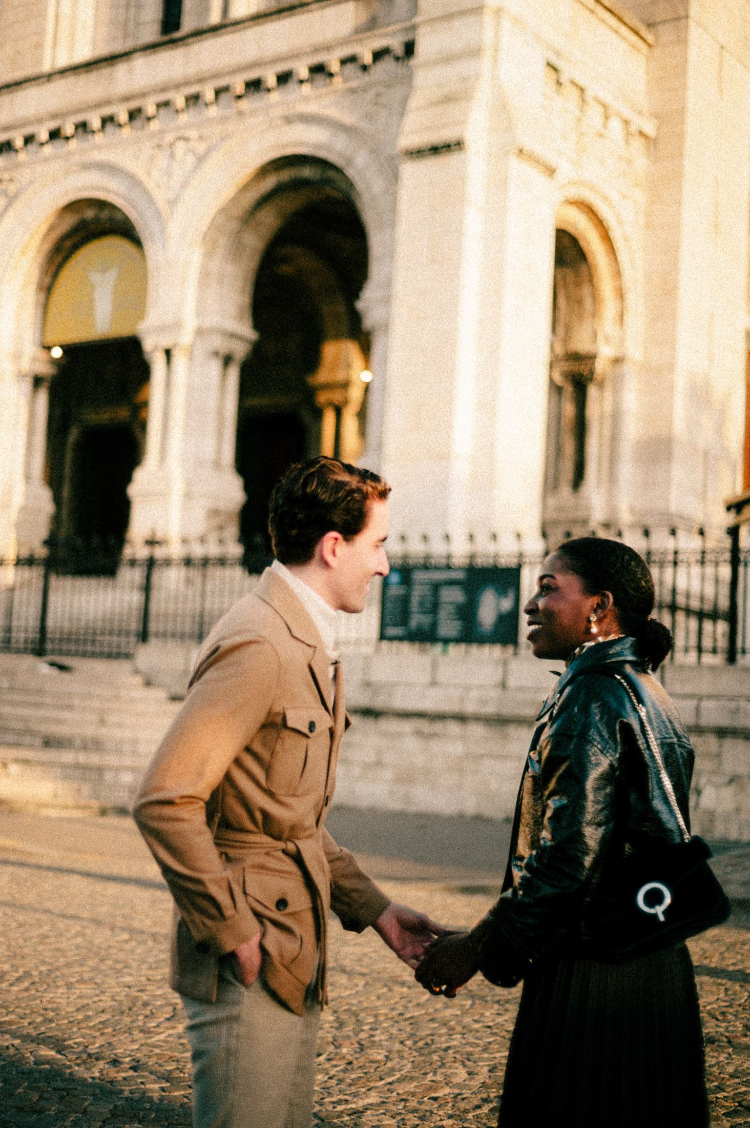 Two people share a playful moment in front of an ornate stone building with arched columns.