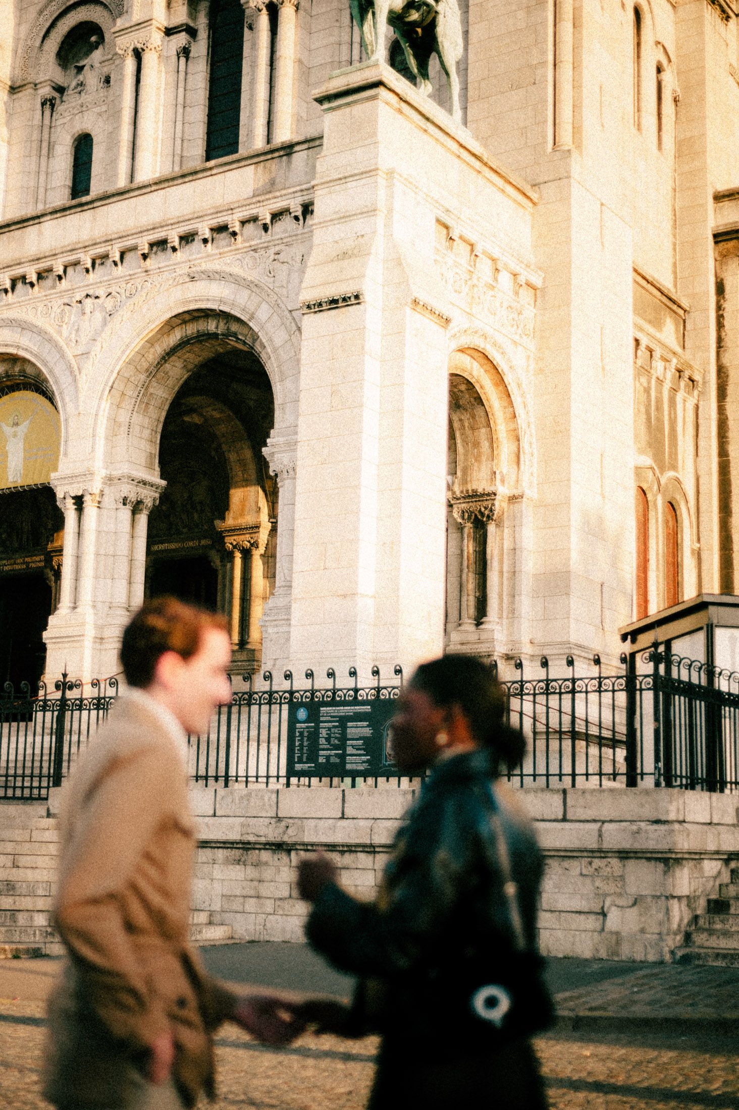 People walking past the ornate stone facade of Sacre-Coeur Basilica in Montmartre, Paris during golden hour.