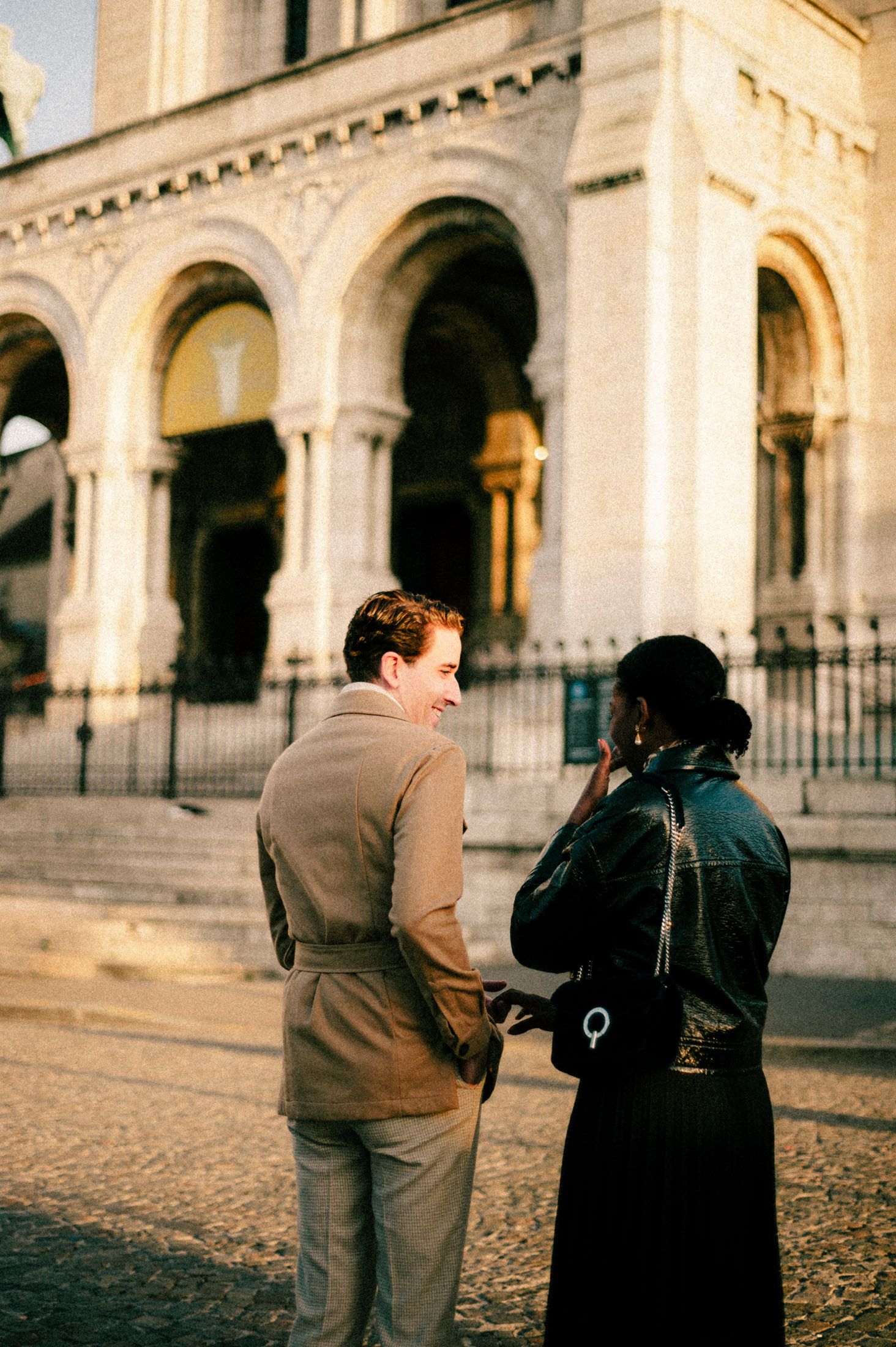 Two people walk together on cobblestone pavement beside an ornate classical building with arched columns.