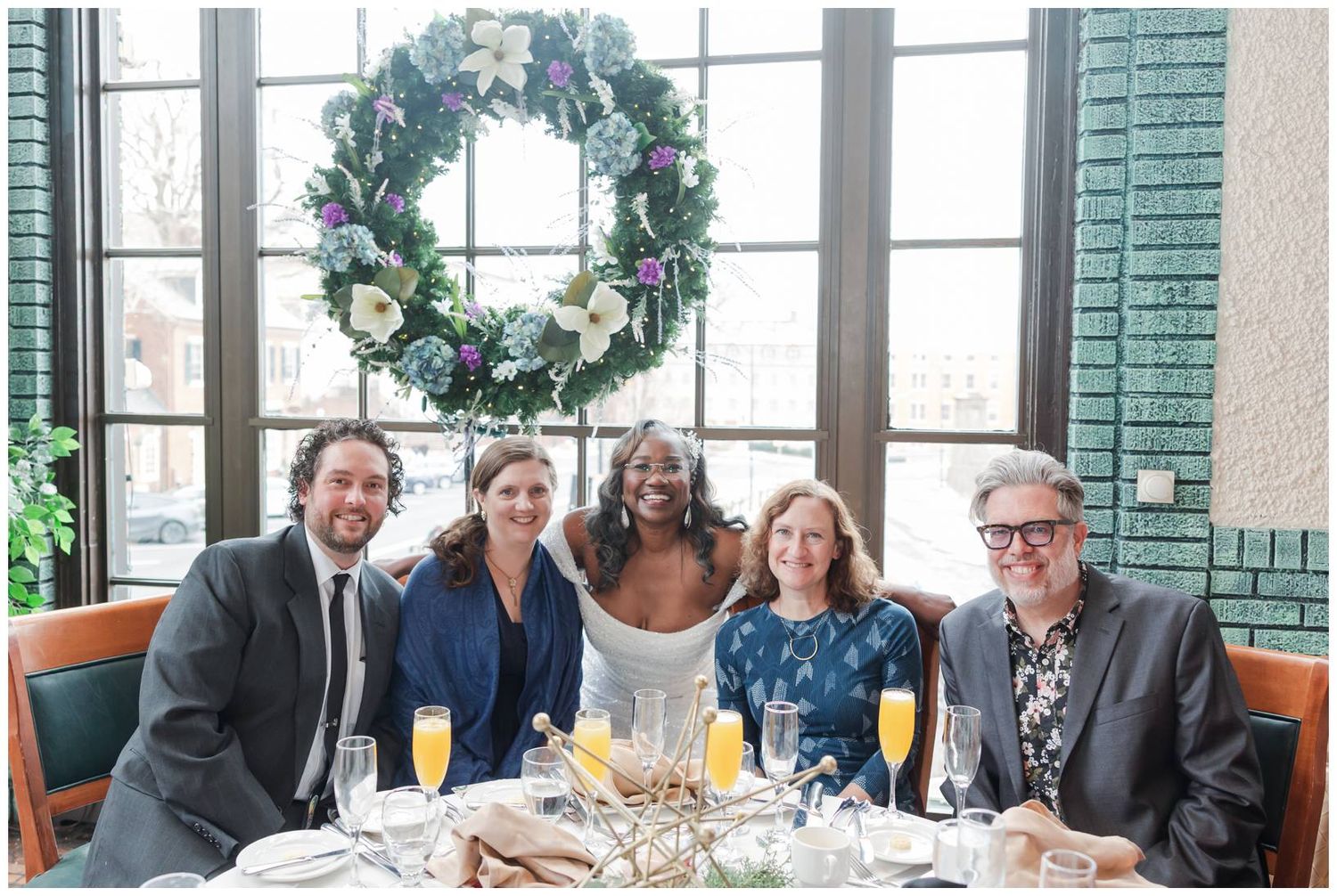 Wedding reception guests seated at tables with holiday decorations and large windows in background.