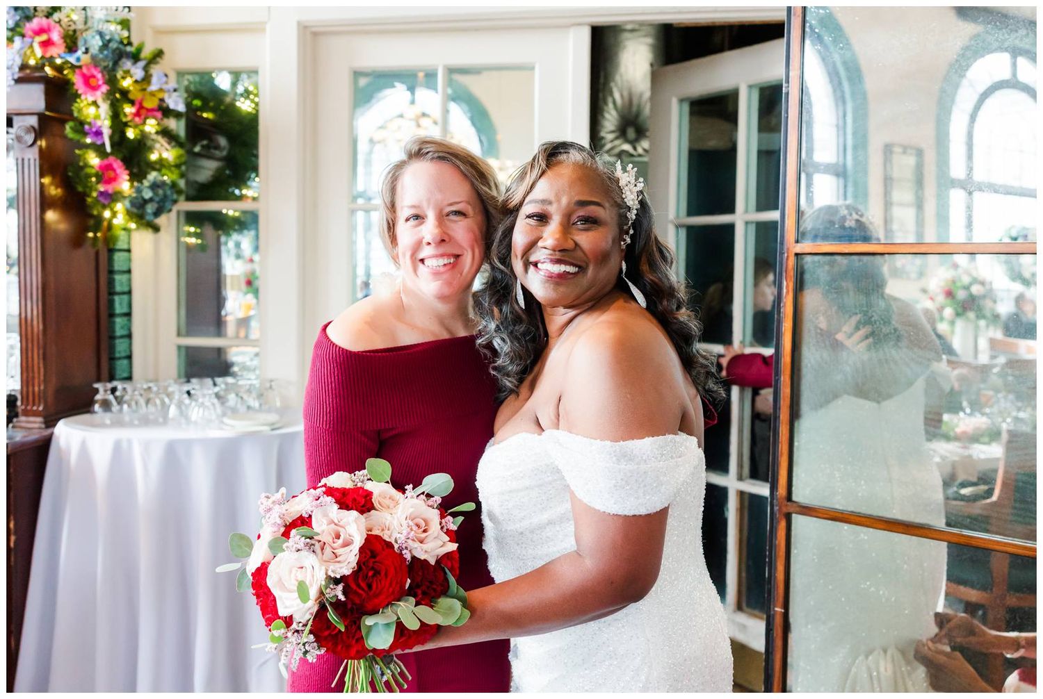 Two people pose together at an elegant wedding celebration with red and white floral bouquets.