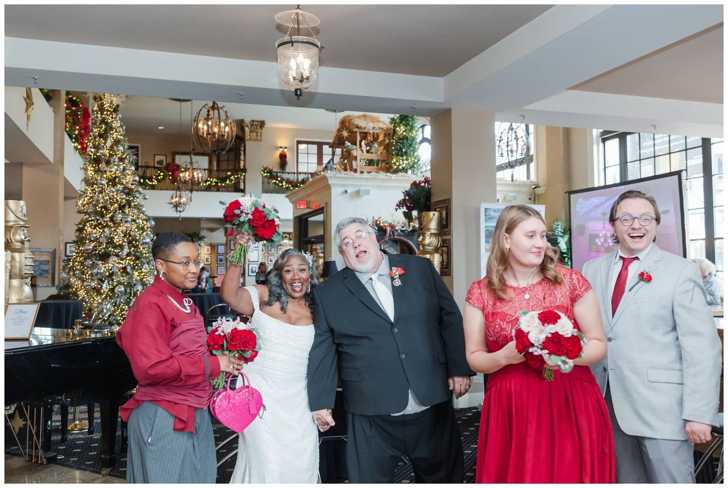 A festive Christmas wedding party stands together in a decorated room with a decorated tree and piano in the background.