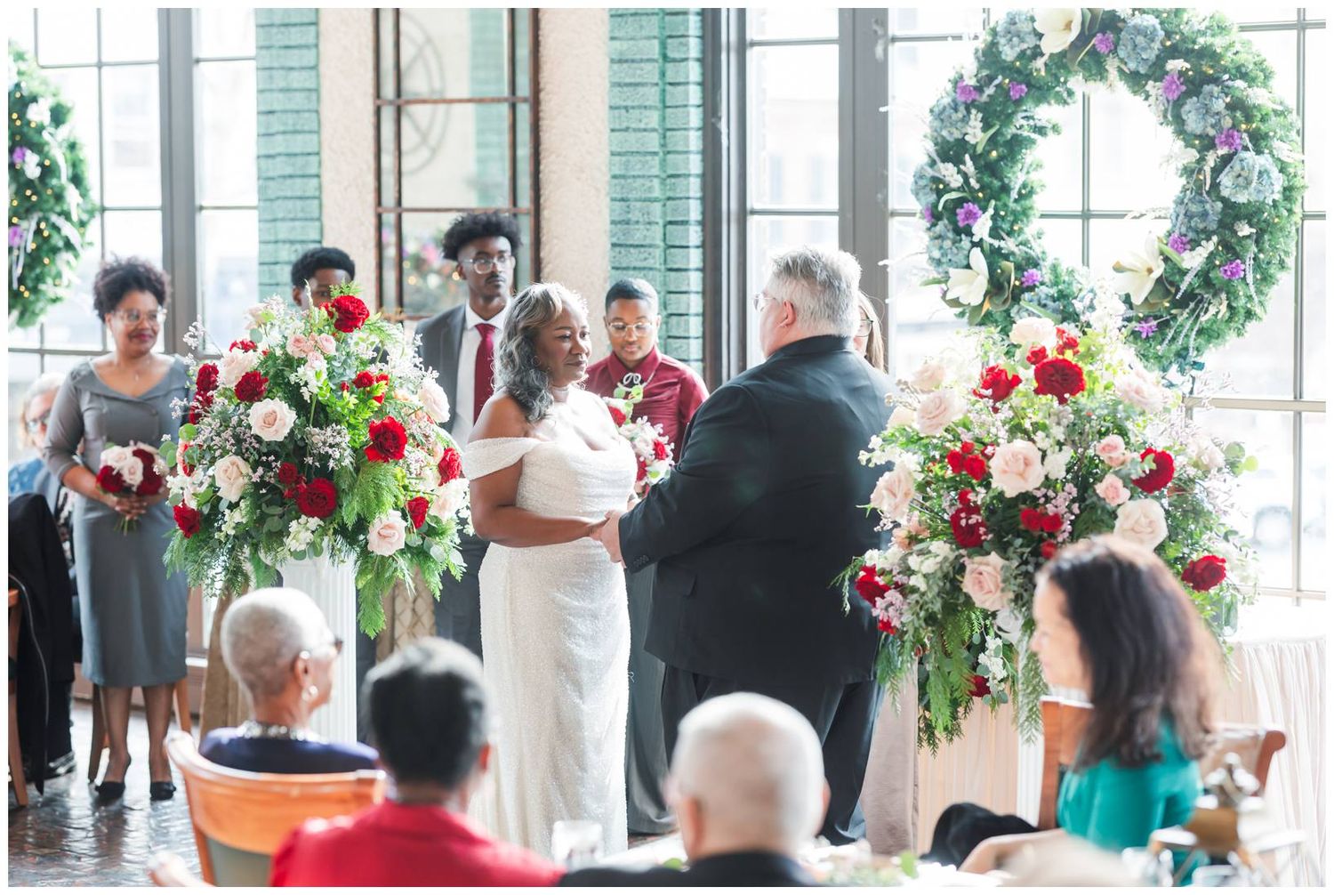 Wedding ceremony takes place under a beautiful floral arch with red and white flowers in an elegant indoor venue.