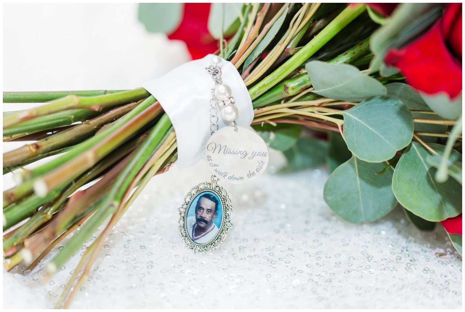 A delicate silver pendant necklace with blue gemstone displayed among red roses and eucalyptus leaves on white lace.