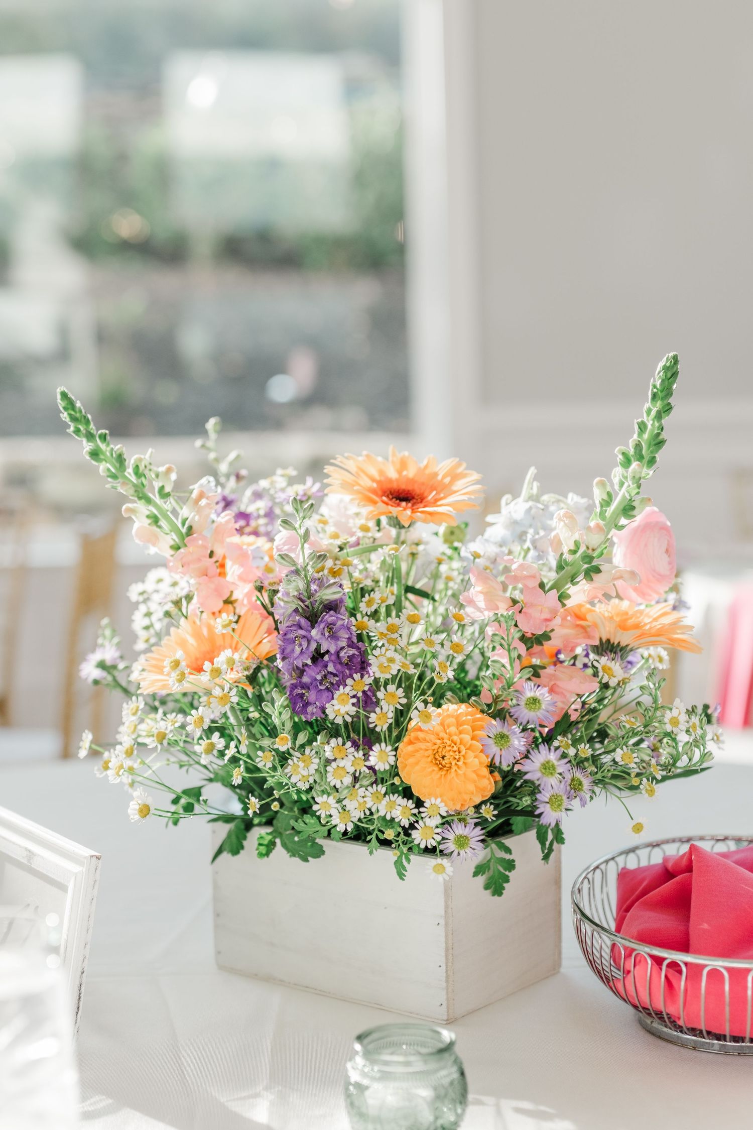Colorful spring wedding centerpiece with gerbera daisies, snapdragons, and wildflowers in a white box vase.