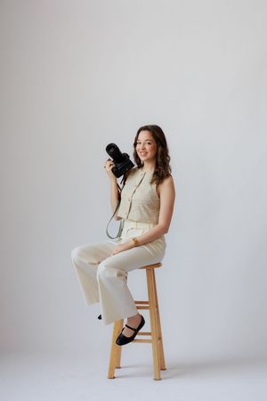 A photographer in neutral toned outfit sits on wooden stool holding camera against white background.