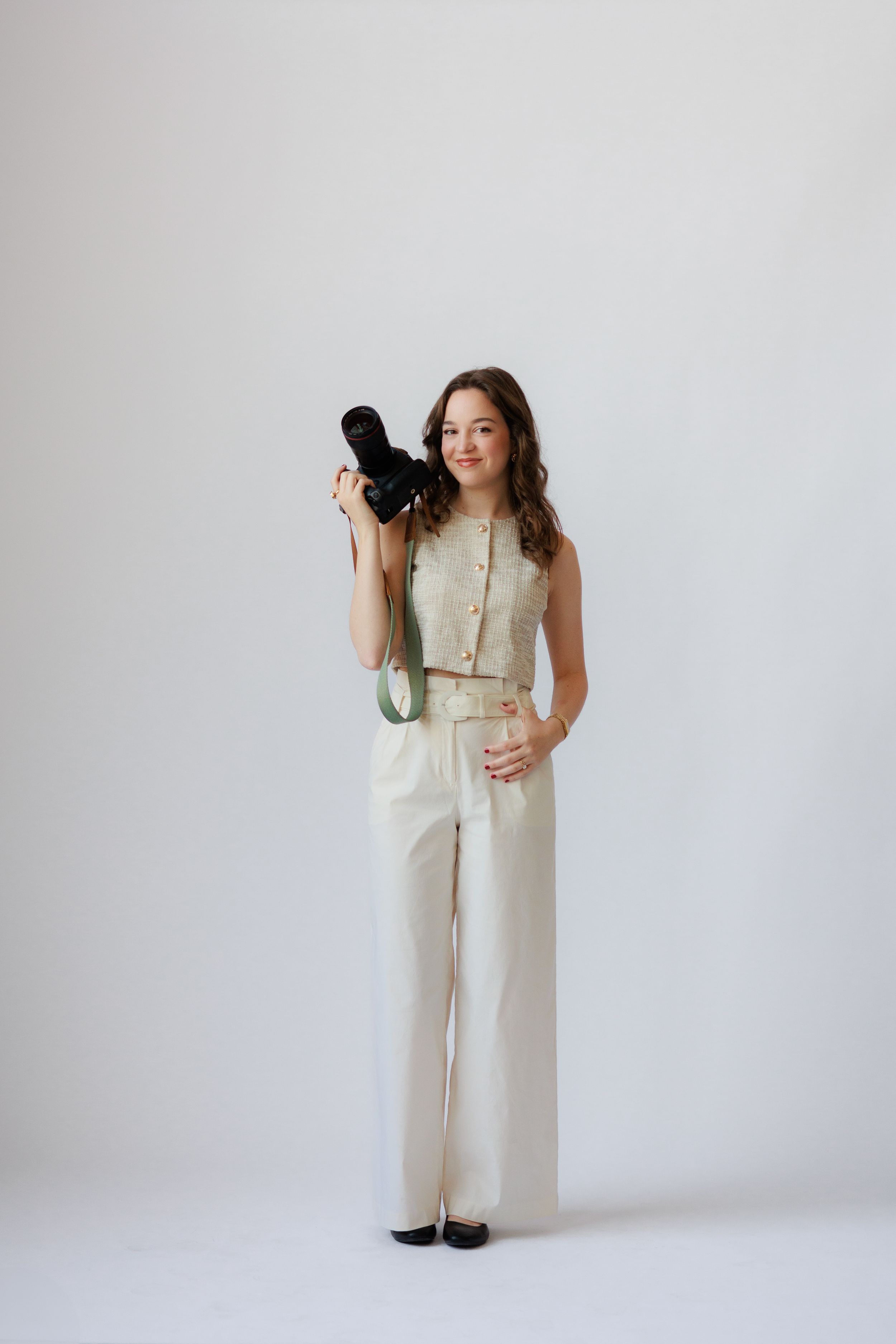 A person in a cream colored sleeveless jumpsuit holds a camera while posing against a white background.