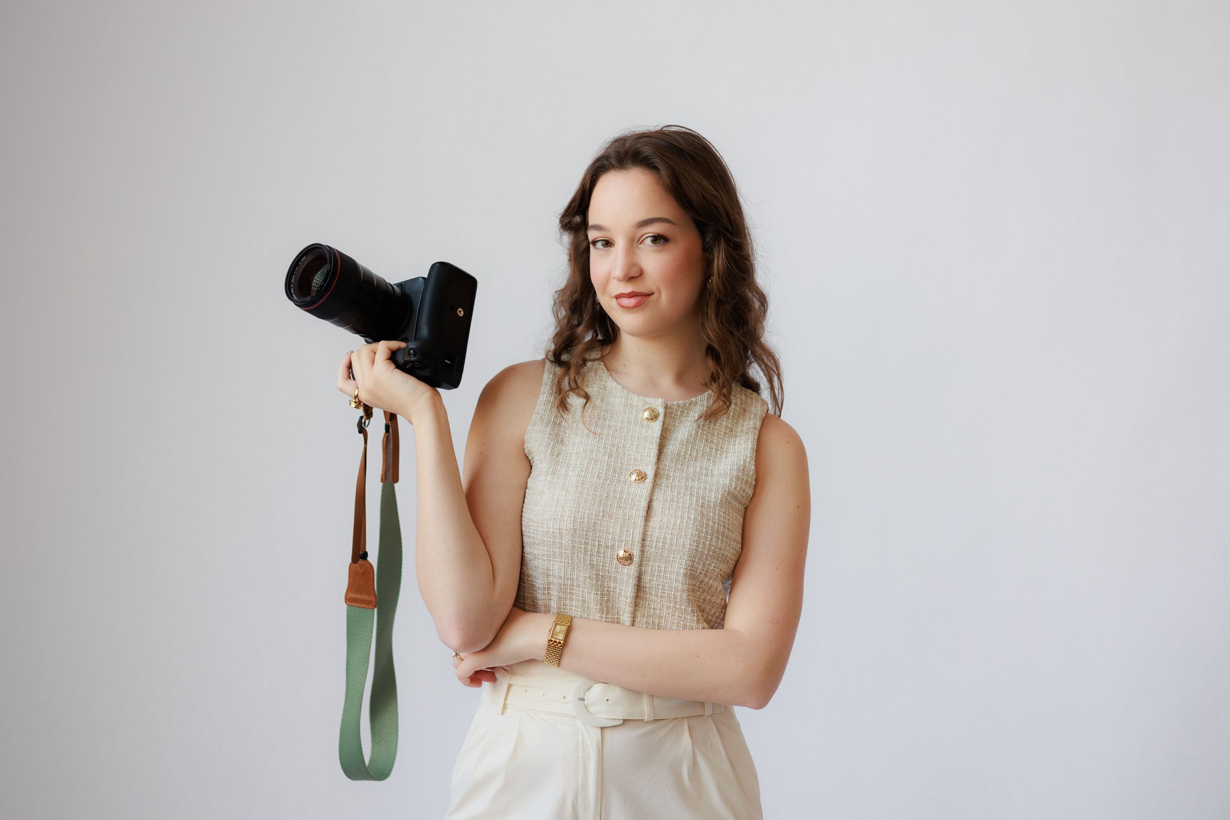 Professional photographer in beige sleeveless dress holds DSLR camera against white studio backdrop.