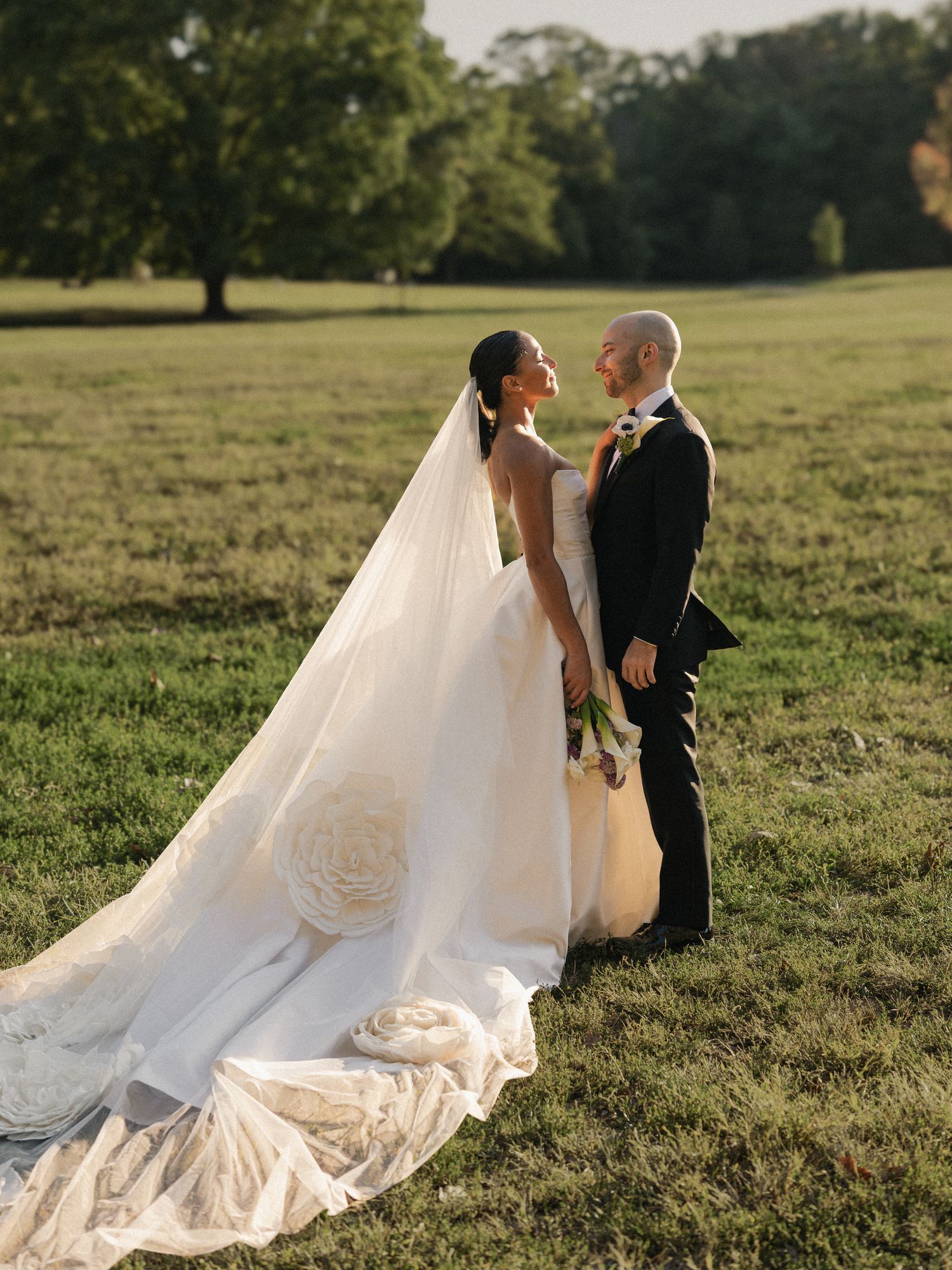 A bride in a flowing white wedding gown and veil walks along a curved stone path through a garden setting.