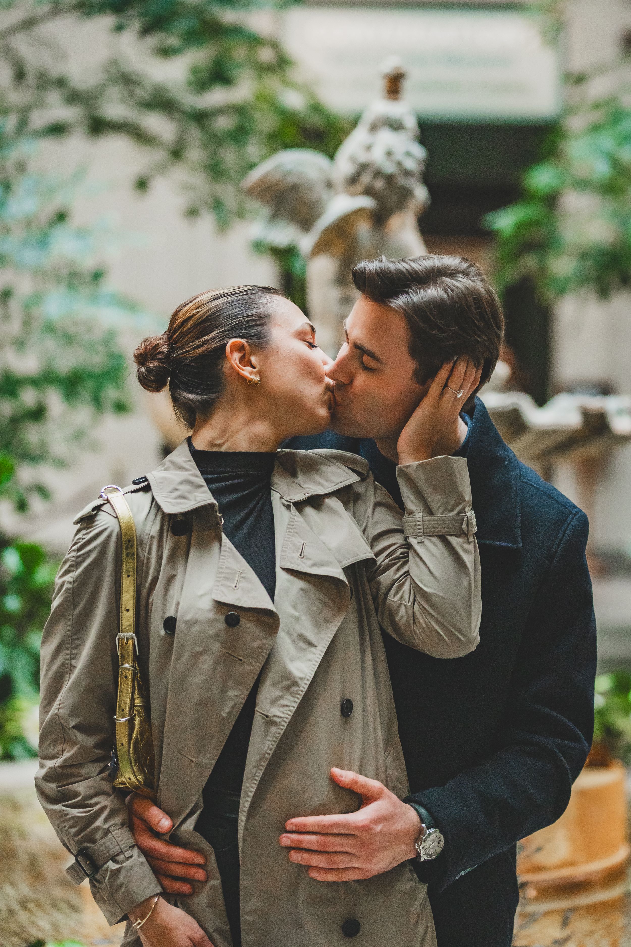 A romantic couple sharing an intimate kiss while wearing a beige trench coat and navy blazer in a garden setting.