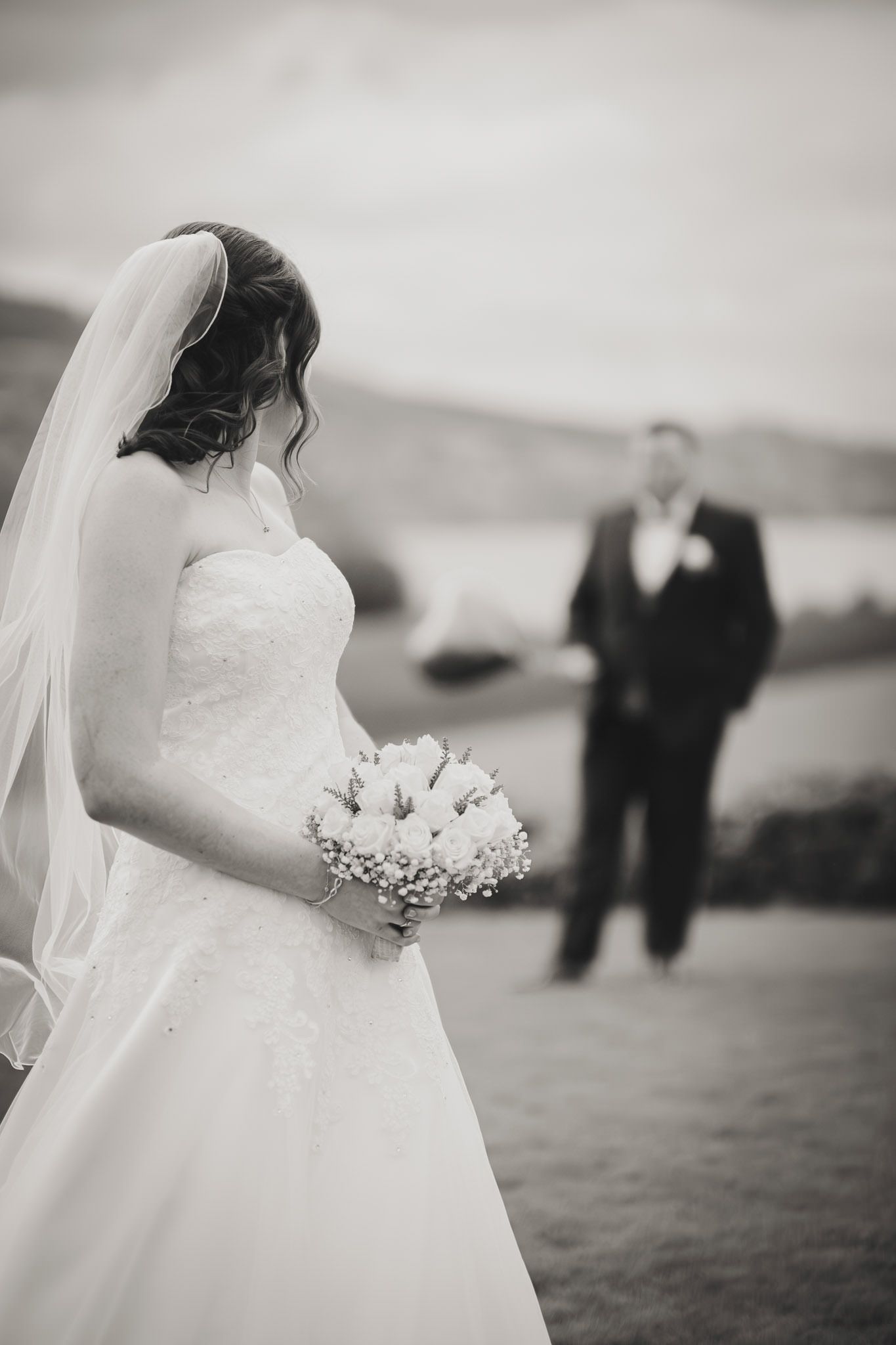A bride in a white wedding gown holds a bouquet while standing on a hilltop.